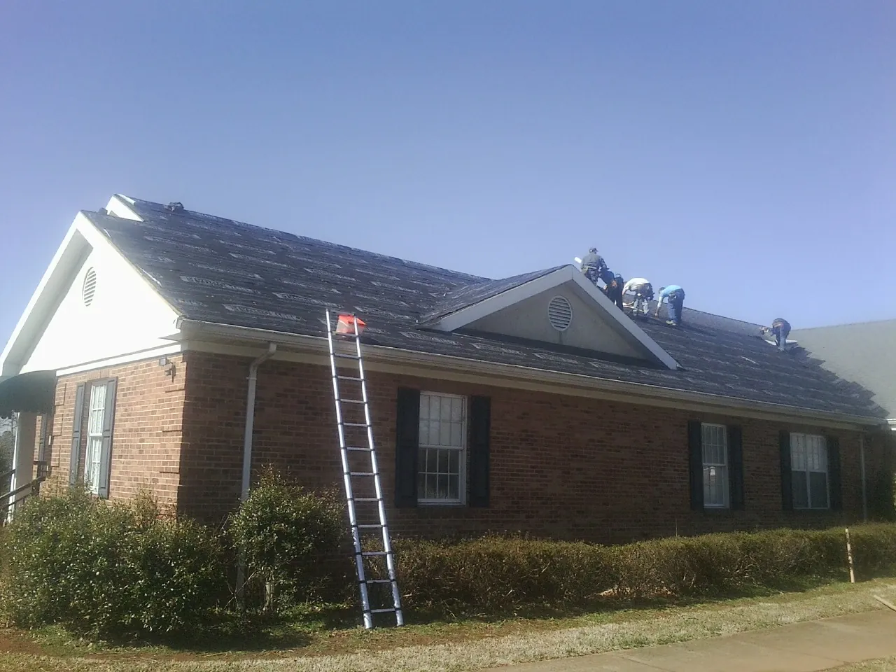 Crew tearing shingles off a brick ranch roof with a ladder set — Metal Roof Repair work by our crew in Wood Lake