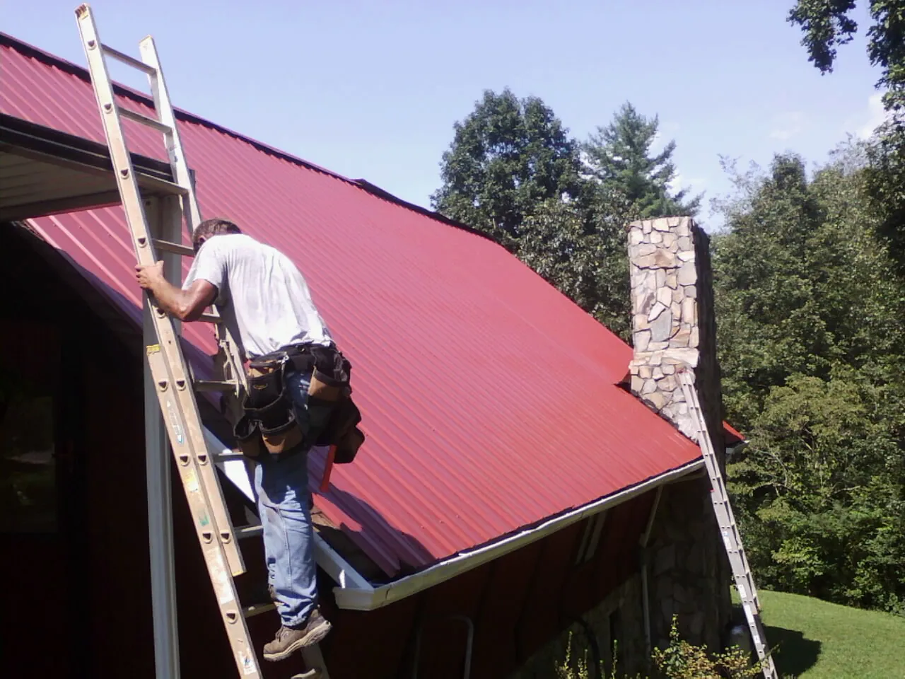 Roofer on a ladder beside a red metal roof and stone chimney — Soffit & Fascia Repair by Roofing Brothers in Wood Lake